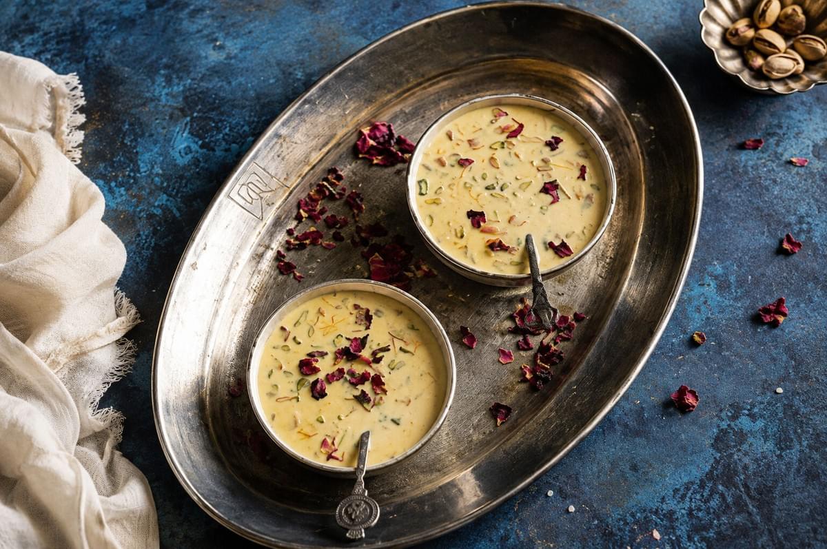 Two silver bowls of kheer topped with pistachios and dried rose petals served on a metal tray.