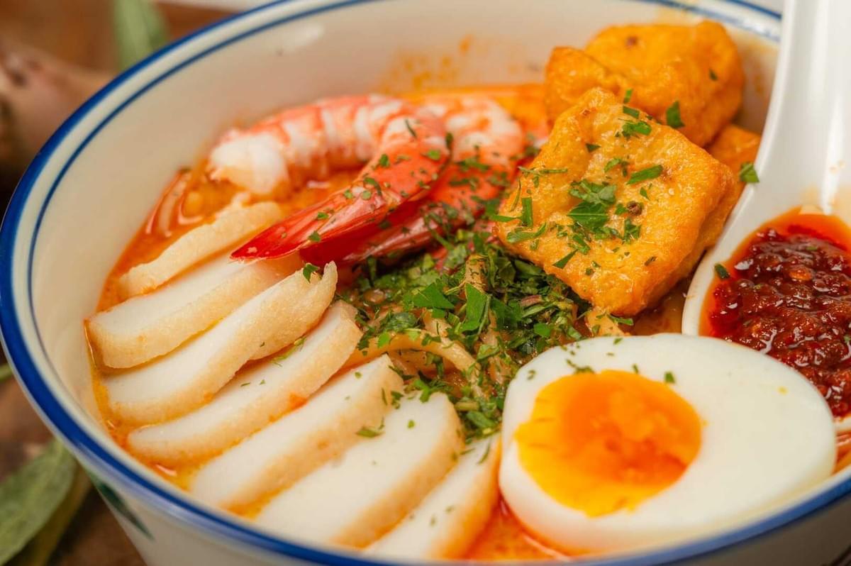 A bowl of Laksa featuring shrimp, eggs, and various colorful ingredients in a rich broth.