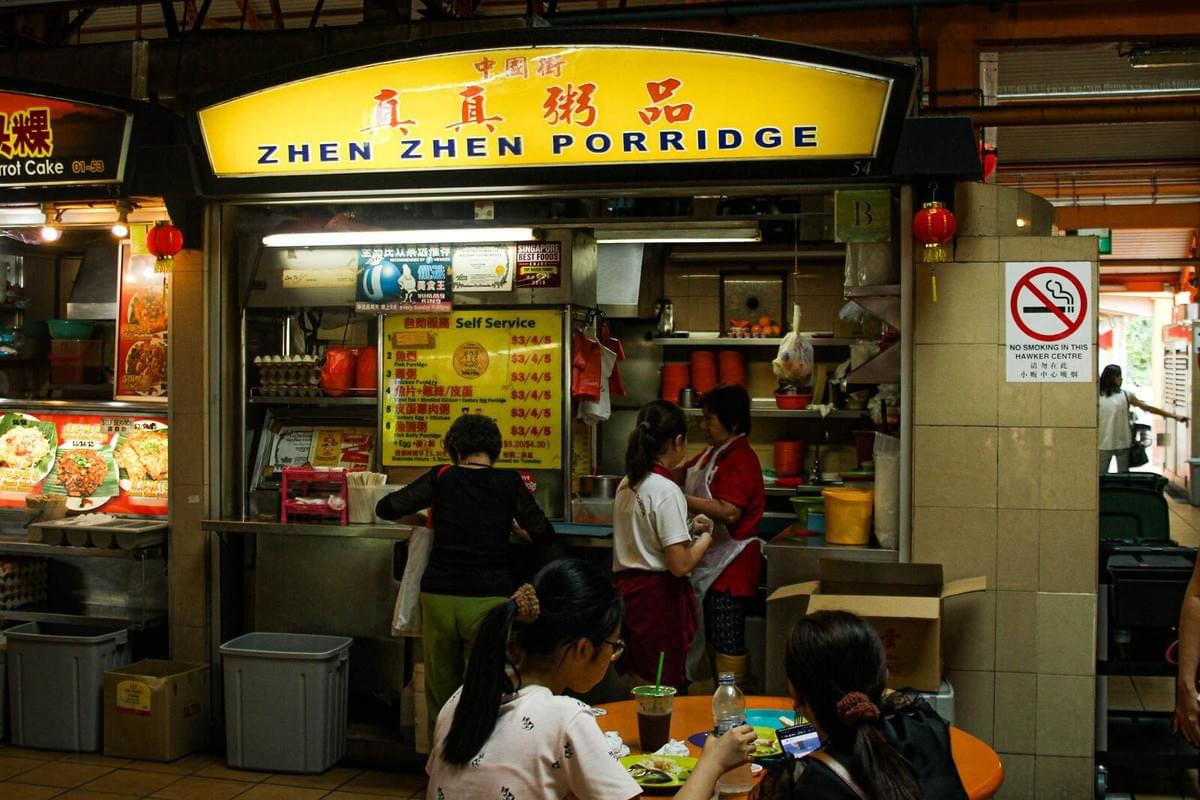 A brightly lit hawker stall named "Zhen Zhen Porridge" serves customers in a busy Singaporean food center. People are seen ordering food and dining at a nearby orange table, surrounded by various signs and kitchen equipment.