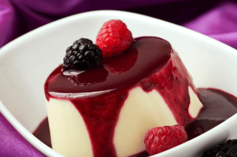 A smooth, unmolded Panna Cotta in a white bowl, generously covered in a dark purple berry sauce and topped with fresh blackberries and raspberries.