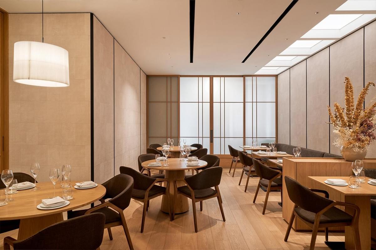 An elegant, modern restaurant dining room features light wood tables, dark modern armchairs, and neutral beige walls. The space is illuminated by a drum pendant light on the left, skylights along the right wall, and translucent Japanese-style shoji sliding screens at the back.