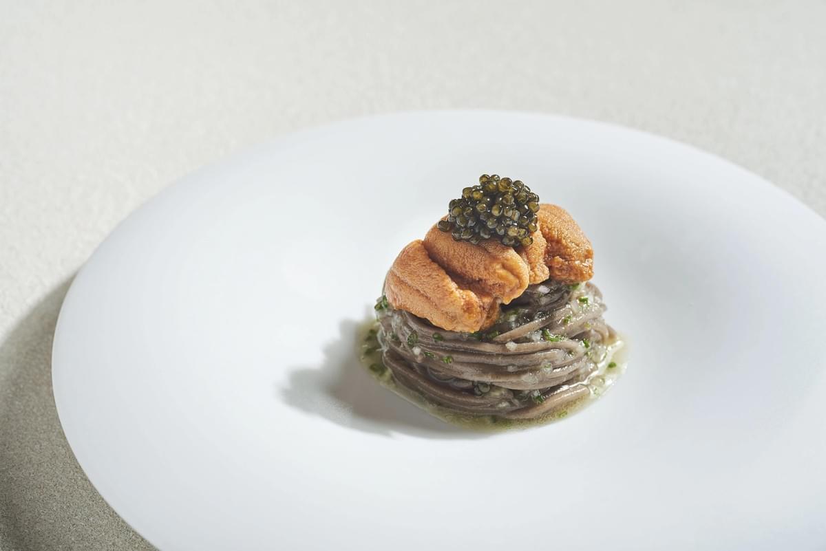 A neatly swirled nest of buckwheat noodles sits in a shallow white plate dressed with oil and fine herbs. The dish is topped with bright orange uni (sea urchin) tongues and a generous mound of glistening black caviar.