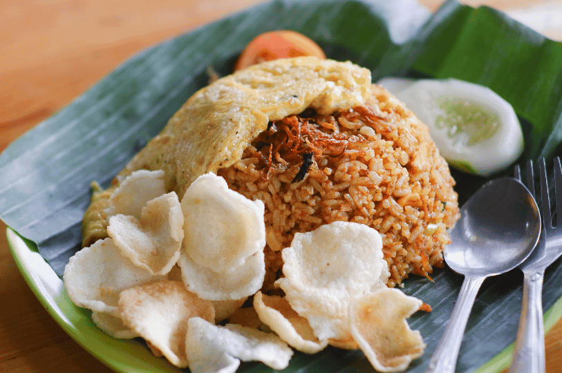 Authentic Nasi Goreng served on a traditional banana leaf, topped with a folded omelet and crispy fried shallots, accompanied by white prawn crackers and fresh tomato slices.