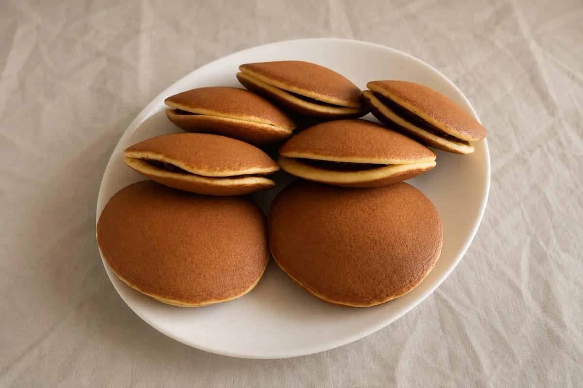 A close-up shot of a pair of hands gently breaking open a fluffy dorayaki pancake, revealing a generous, textured filling of sweet red bean paste inside. In the blurred background, two whole, golden-brown dorayaki sit on a wooden serving board against a muted purple-grey backdrop.