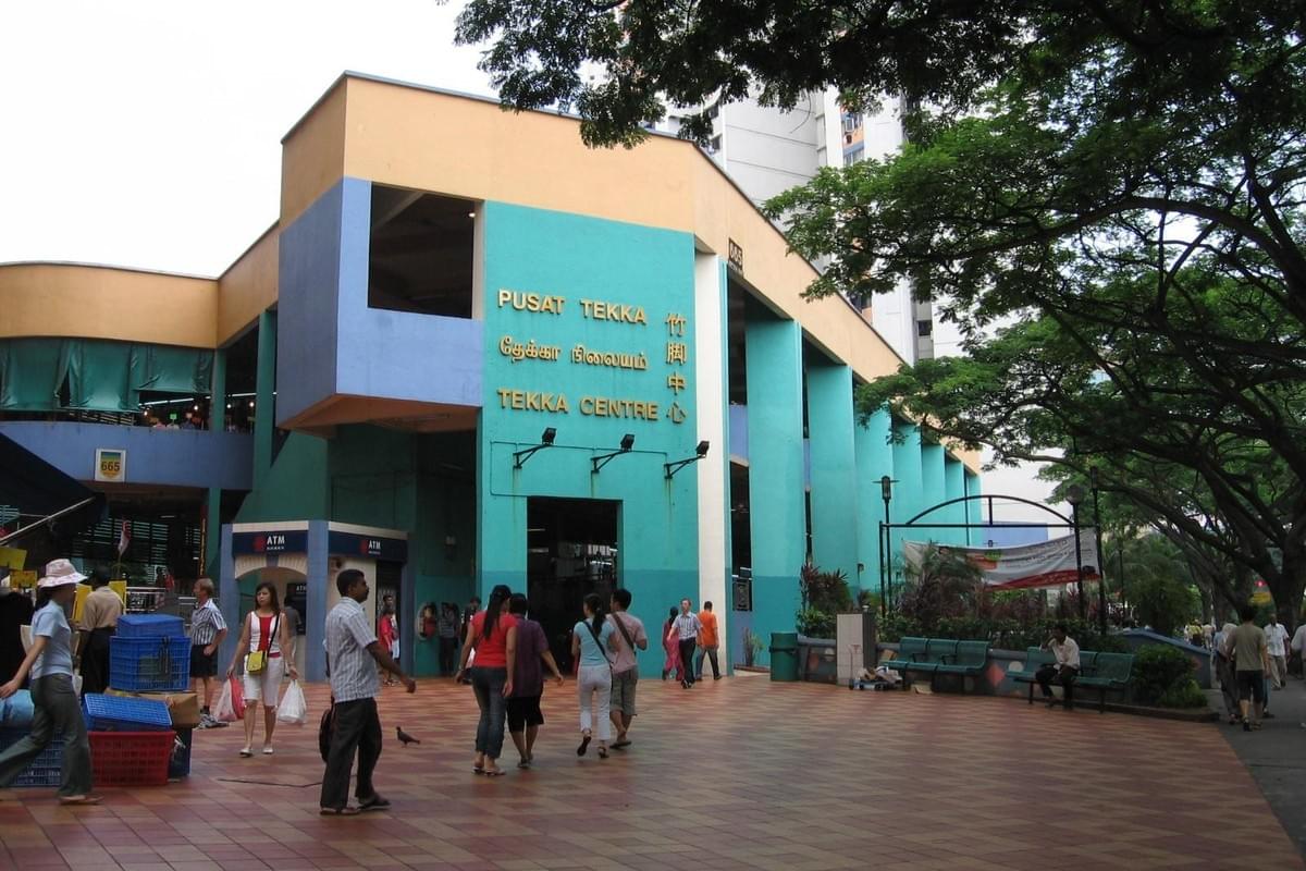 The image shows the exterior of the Tekka Centre in Singapore, a vibrant building with teal, yellow, and blue geometric accents. People are seen walking across a tiled plaza in the foreground under the shade of large, leafy trees.