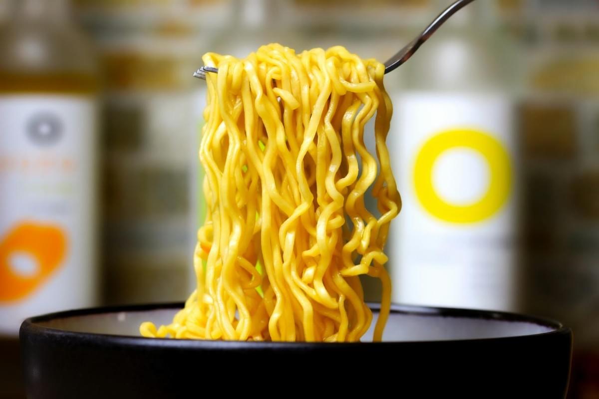 A close-up shot shows a large cluster of wavy, yellow instant noodles being lifted on a fork out of a dark bowl. In the softly blurred background, two bottles with white and yellow labels are visible.