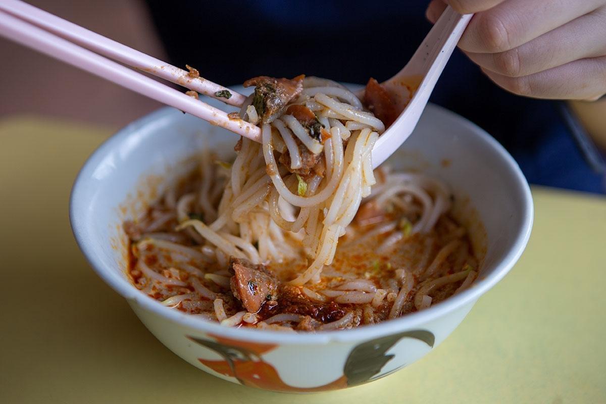 A person uses pink chopsticks and a matching spoon to lift thick white laksa noodles from a flavorful orange broth. The dish is served in a traditional rooster-motif bowl and contains bean sprouts and pieces of cockle.