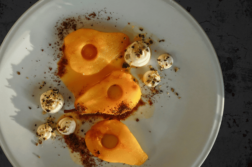 A gourmet plated dessert featuring three poached pear halves drizzled with amber syrup and spiced crumbles, accompanied by dollops of whipped cream or meringue.