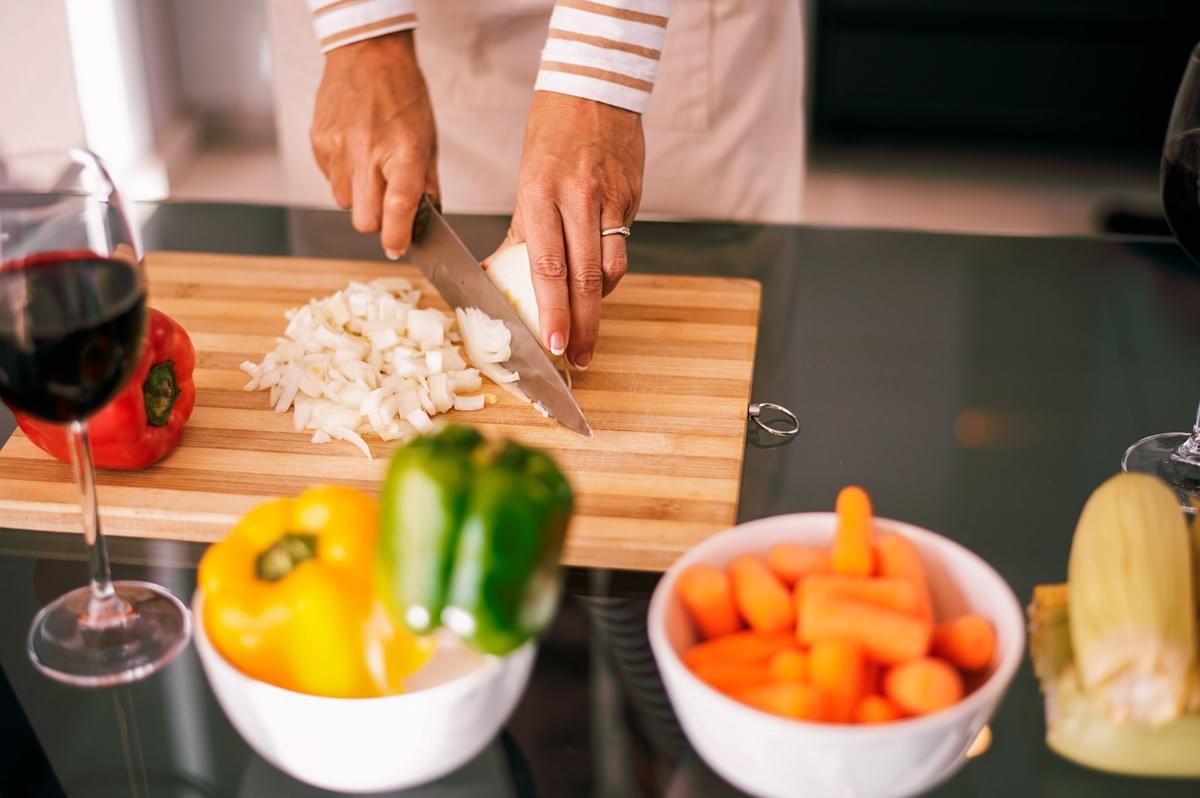 Hands chopping onions on a wooden cutting board surrounded by bell peppers, baby carrots, and a glass of red wine.