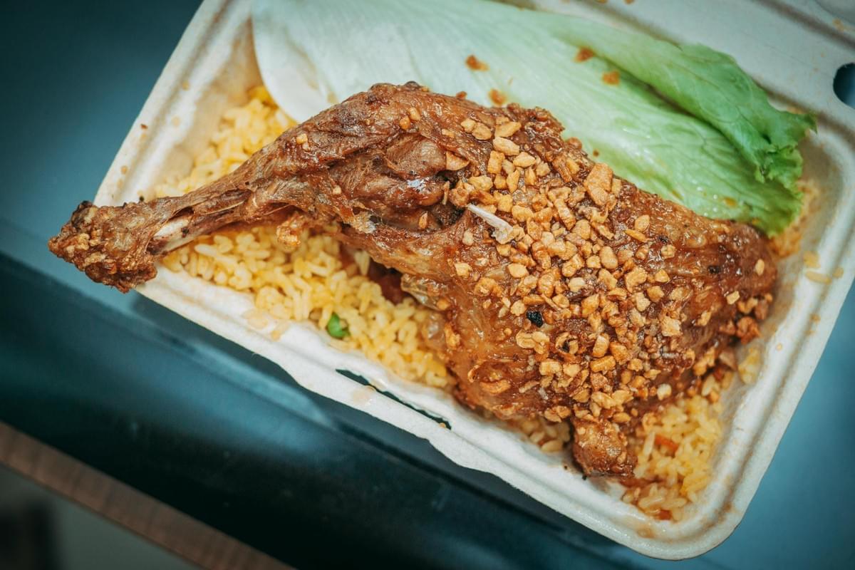 A close-up shot displays a golden-brown fried chicken leg resting on a bed of yellow rice within a white takeout container. The crispy skin is generously topped with fried garlic bits, and a fresh lettuce leaf garnishes the side of the meal.