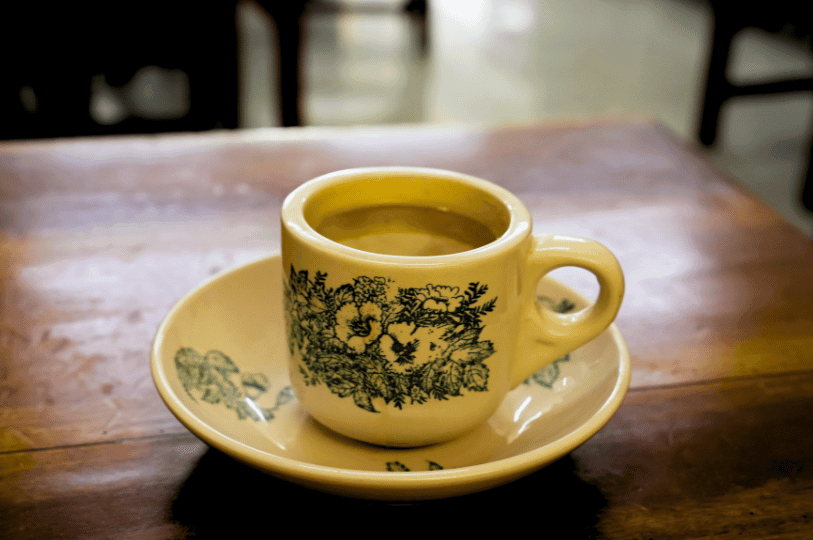 A classic vintage kopitiam cup and saucer decorated with green botanical motifs, resting on a polished wooden table, highlighting the traditional glassware used for Singapore kopi.