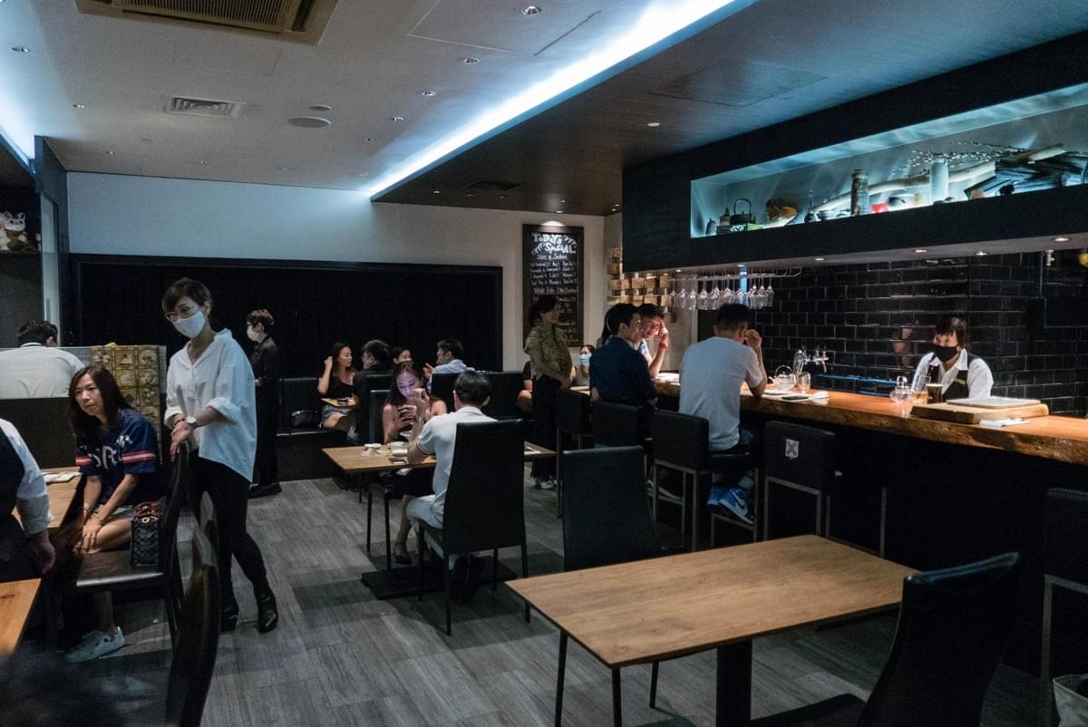 An indoor shot shows customers dining in a modern restaurant with dimly lit, blue-accented ceilings, wooden tables, and dark chairs. Several patrons are seated at a long bar counter on the right where a staff member wearing a face mask stands behind the counter.