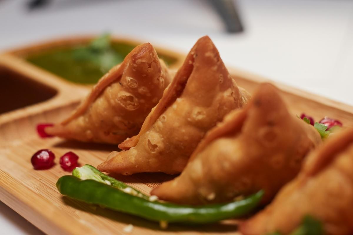 The image shows a close-up, angled view of golden-brown, crispy samosas arranged on a wooden platter. They are accompanied by a fresh green chili, a few pomegranate seeds, and a side of vibrant green chutney.