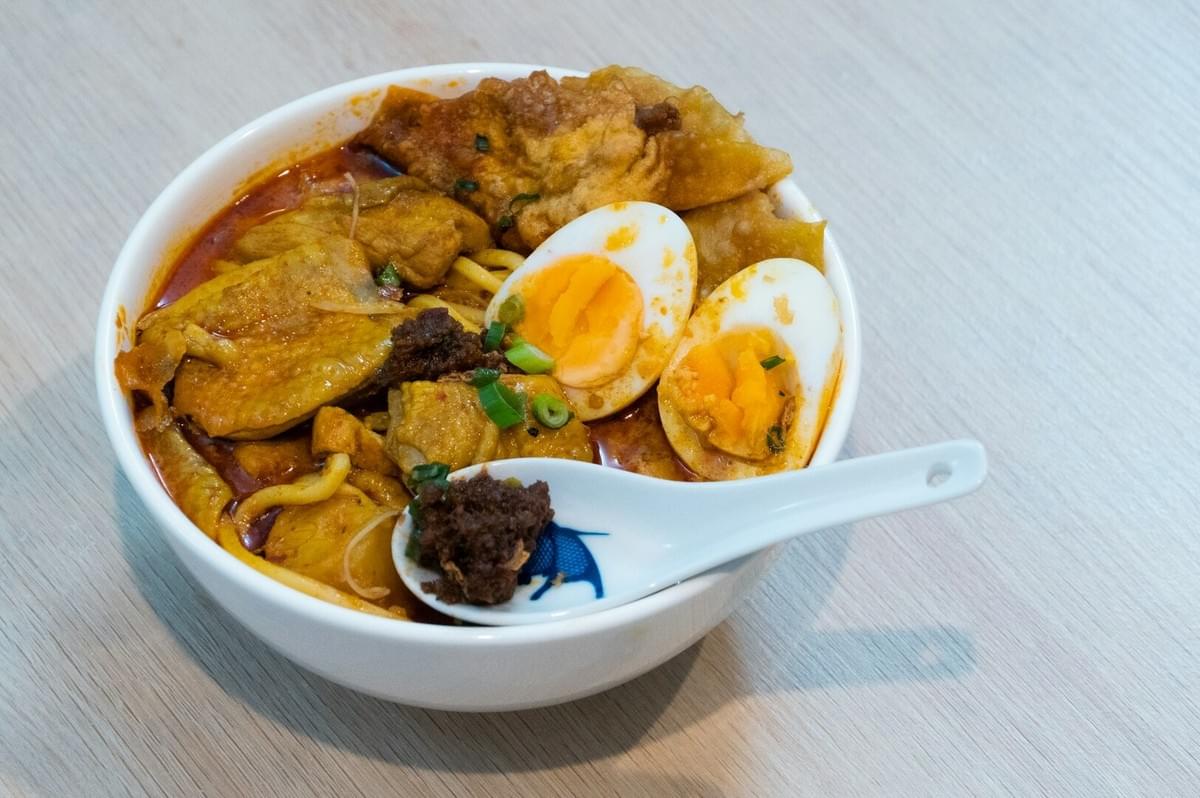 A bowl of Laksa featuring noodles, meat, and eggs in a flavorful broth.