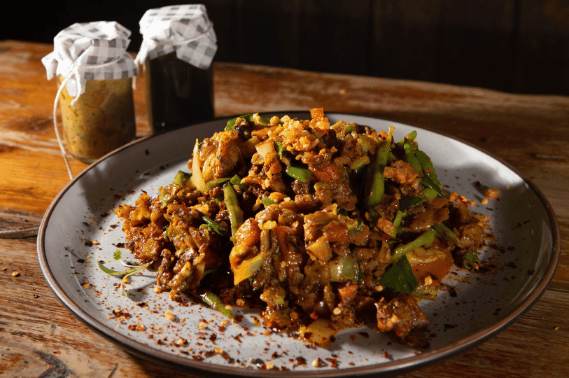 A dark, moody shot of a rich, meat-filled Kottu Roti served on a grey plate, garnished with green chilies and served alongside jars of spicy condiments.