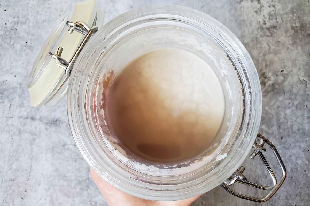 An overhead view of a neglected sourdough starter in a glass jar, showing a layer of thin, dark liquid, known as hooch, floating on the surface.