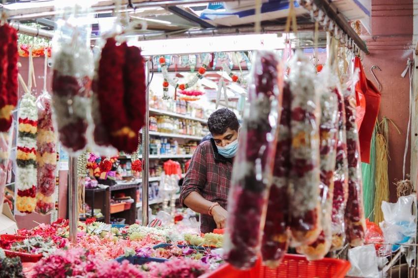 A man wearing a plaid shirt and a protective face mask speaks on a mobile phone while standing behind a market stall abundantly stocked with colorful flower garlands and loose petals. The scene is visually framed by blurred, plastic-wrapped garlands hanging in the foreground