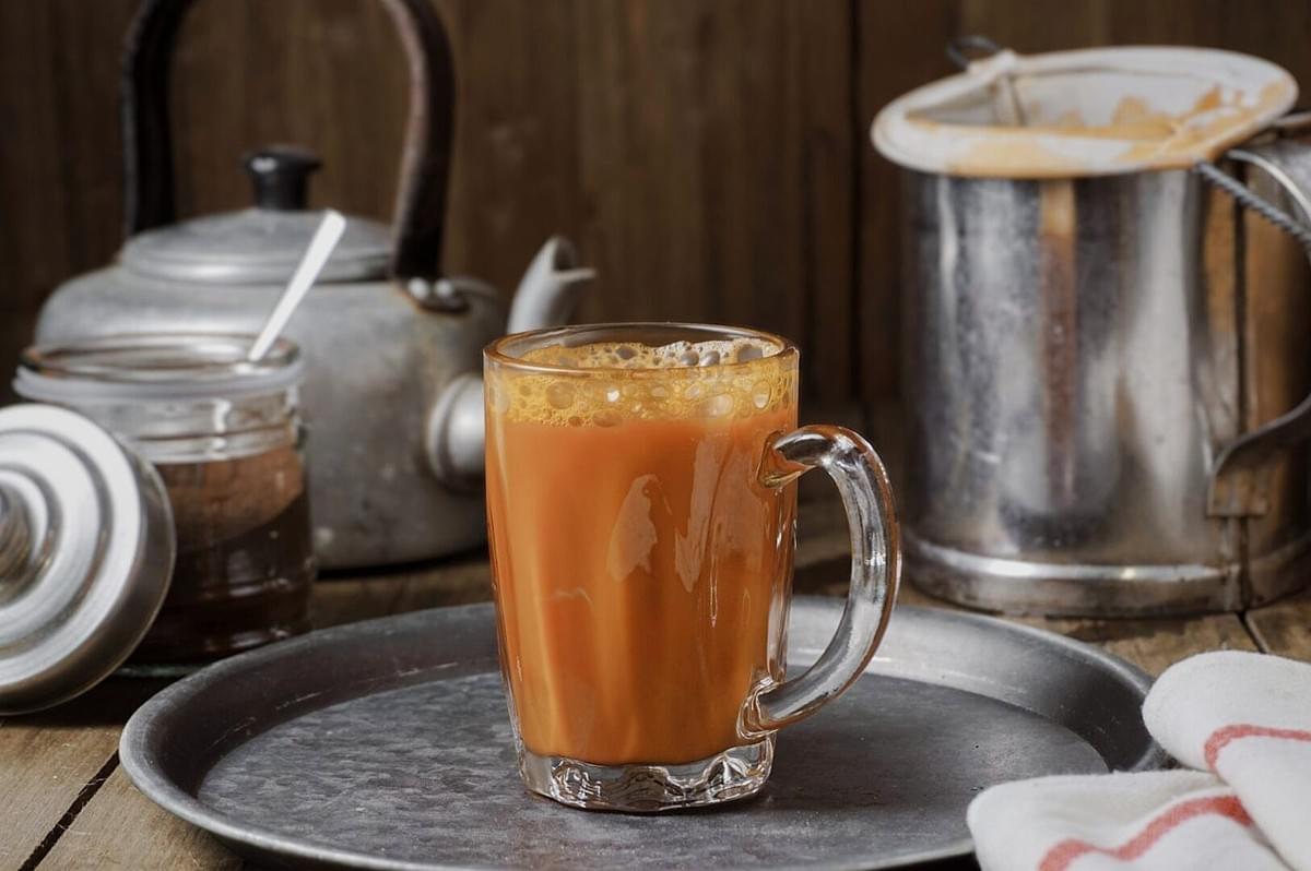 A glass of tea on a plate next to a silver pot, showcasing Teh Tarik, a traditional Singaporean beverage.