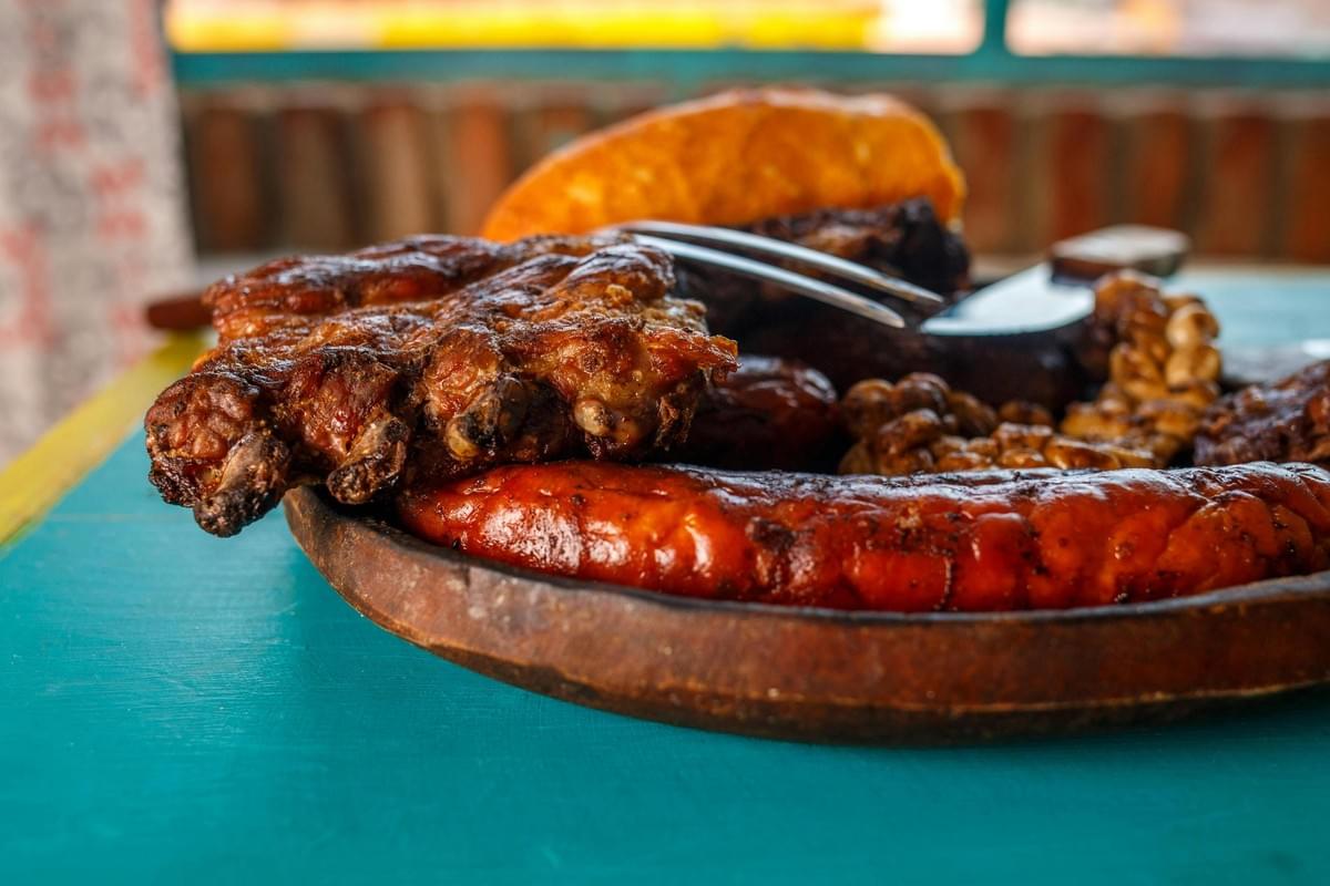 A wooden platter overflowing with grilled meats, including a thick sausage and charred ribs, sits prominently on a bright turquoise surface. In the background, a warm orange roll and various side dishes complete the hearty meal under soft, natural light.