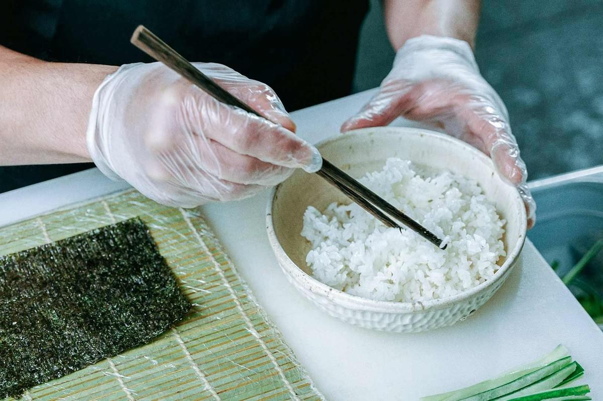 Sushi preparation close-up: gloved hands, bowl of rice, nori on a plastic-covered makisu, and cucumber filling ready to roll.