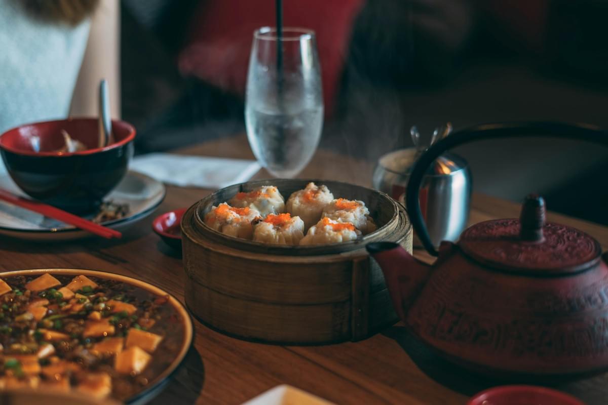 A steaming bamboo basket filled with open-faced shumai dumplings topped with orange roe sits at the center of a restaurant table. Surrounding the basket is a red cast-iron teapot, a plate of mapo tofu, and a glass of iced water.