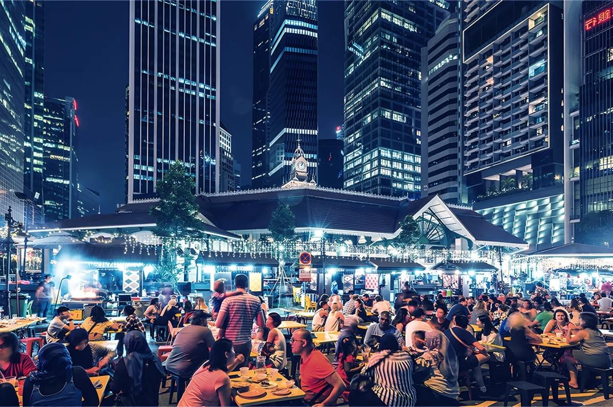 Eye-level wide-angle night photograph of a crowded outdoor hawker center with diners seated at communal tables, illuminated food stalls in the foreground, and modern skyscrapers rising in the background, capturing a vibrant city street food scene.