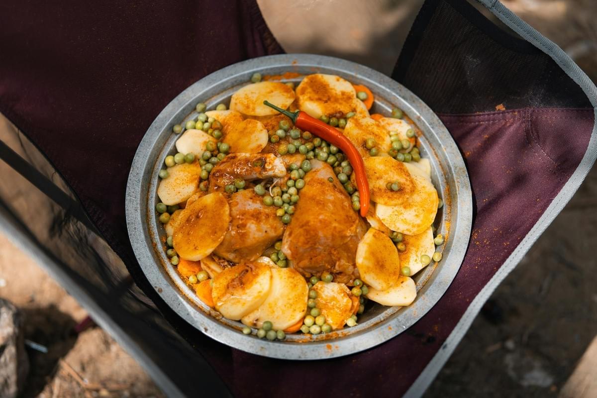A round metal tray is filled with raw ingredients including chicken pieces, sliced potatoes, and green peas, all generously dusted with orange spices. A whole red chili pepper sits atop the arrangement, which is placed inside the seat of a portable camping chair.