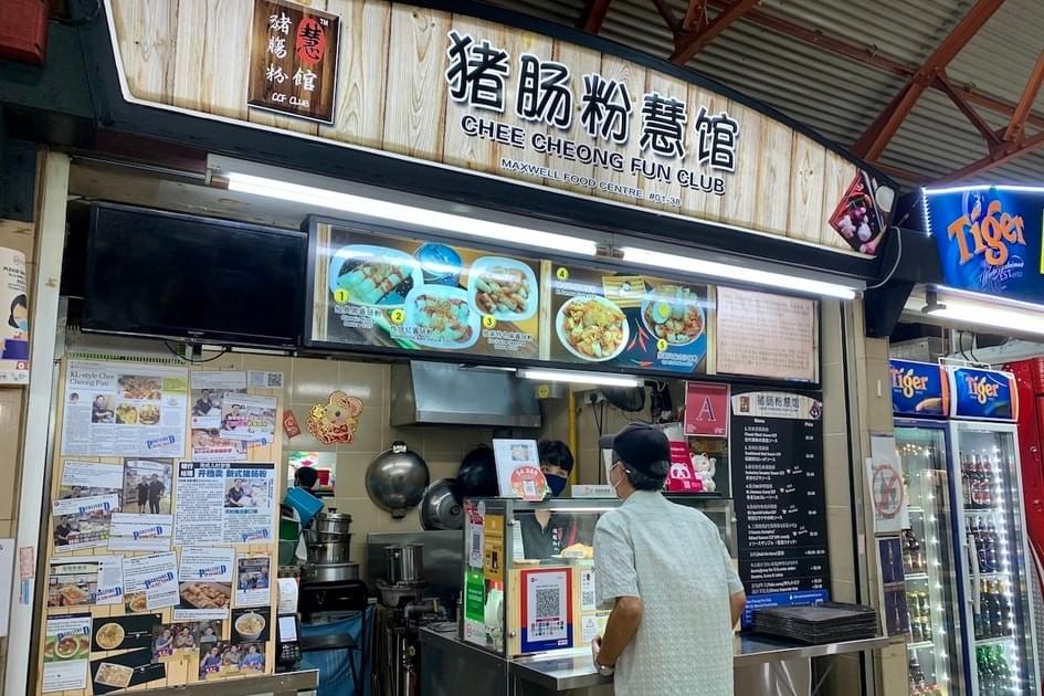 The image shows the Chee Cheong Fun Club stall at Maxwell Food Centre, featuring a rustic wooden-style sign and a menu with numbered photos of various rice noodle roll dishes. A customer in a grey shirt and black cap stands at the counter, which is flanked by a wall of newspaper clippings and a refrigerator stocked with Tiger Beer.