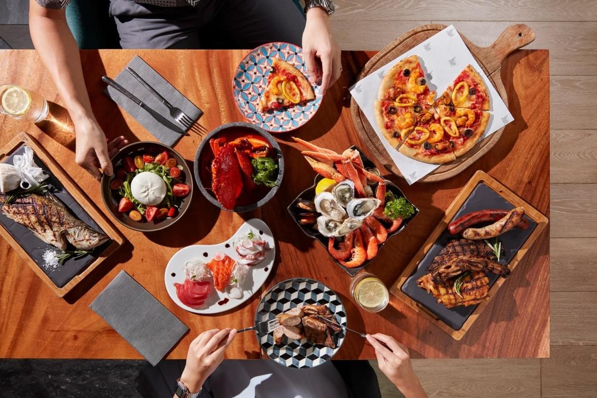An overhead view of a wooden dining table filled with a rich spread including seafood platters, fresh sashimi, pizza, grilled meats, and a caprese salad. Two diners are seated across from each other, reaching for dishes and preparing to enjoy the meal.