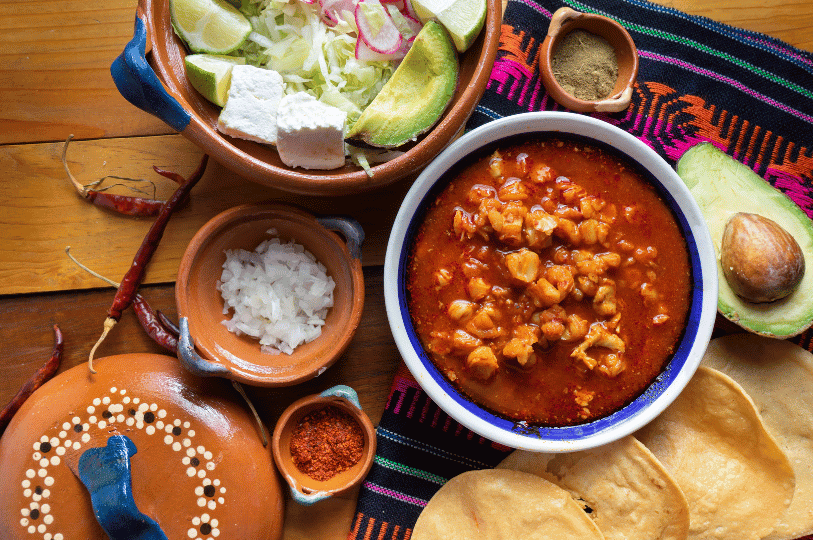 A festive overhead spread featuring a Pozole Rojo surrounded by various toppings including queso fresco, avocado, diced onions, and crispy tostadas.