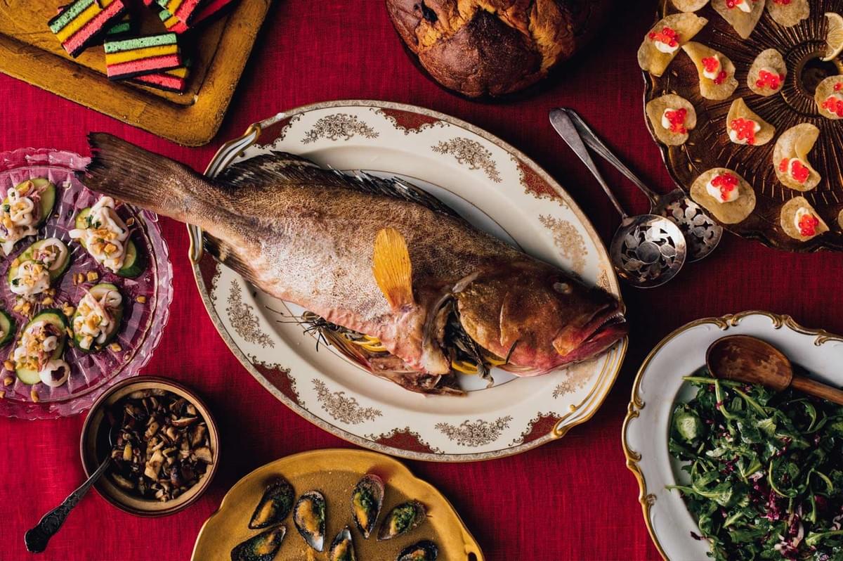 Assorted holiday seafood feast on a red tablecloth featuring a whole cooked fish, salads, appetizers, pastries, and colorful layered desserts.
