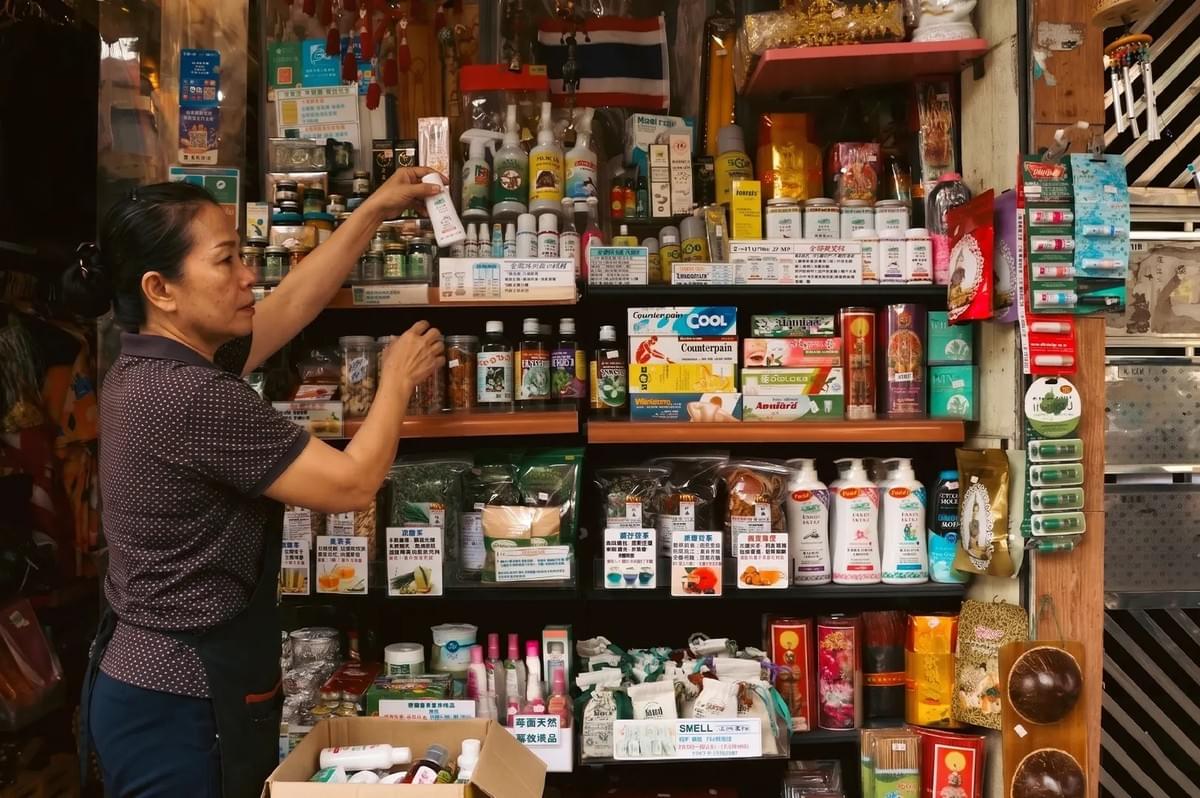 A woman organizing shelves stocked with traditional herbal remedies, balms, and health supplements in a small apothecary or medicinal shop.