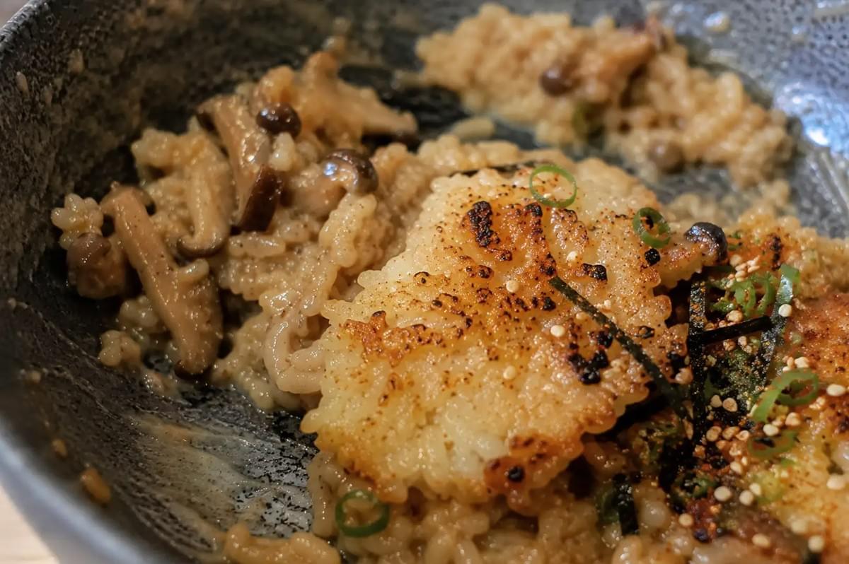 An extreme close-up macro view inside a ceramic dish showing glossy, broth-infused rice cooked with brown shimeji mushrooms, topped with a piece of pan-seared crispy rice featuring charred golden edges, sprinkled with ringed green onions, toasted sesame seeds, chili powder, and fine nori strips.