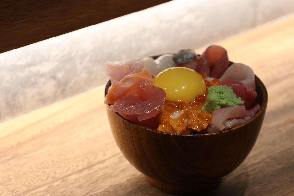 A wooden bowl rests on a light wood counter, filled with a colorful Japanese kaisen don seafood rice bowl. The dish is topped with an raw egg yolk in the center, surrounded by fresh sashimi, sea urchin, salmon roe, and a dollop of green wasabi.