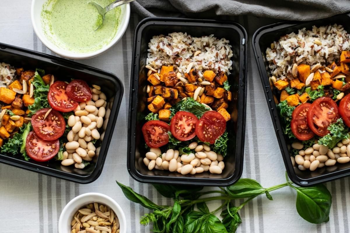 Three black meal prep containers are arranged on a striped tablecloth, each filled with wild rice, roasted sweet potatoes, white beans, kale, and sliced tomatoes. A small bowl of green sauce and a dish of slivered almonds sit alongside fresh basil leaves to complete the healthy spread.