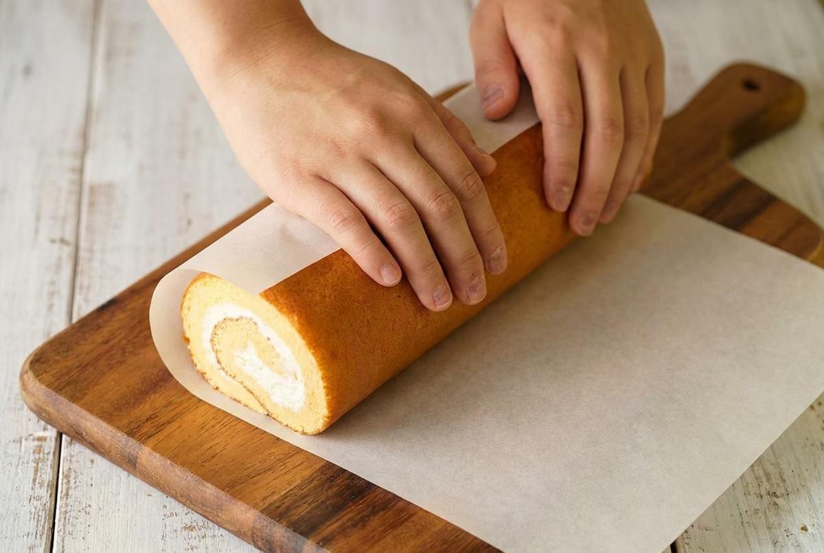 Hands rolling a cream-filled swiss roll cake tightly using parchment paper on a wooden cutting board.
