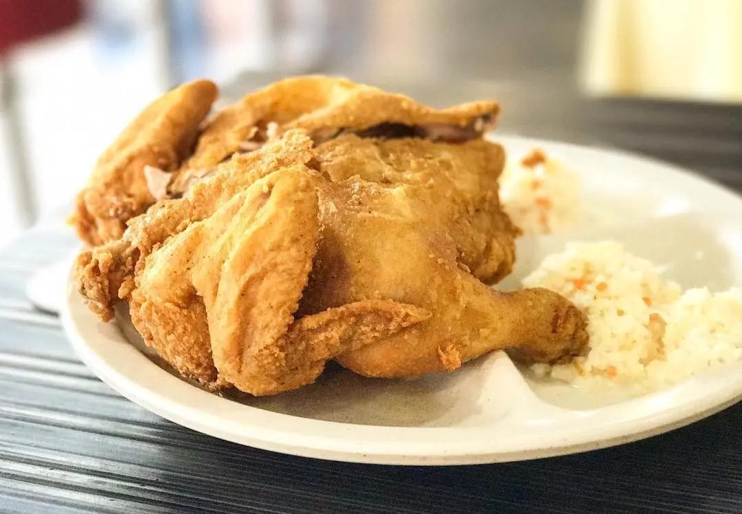 A golden-brown fried half chicken is served on a white divided plate alongside a small portion of coleslaw. The chicken looks crispy and is the main focus against a softly blurred background.