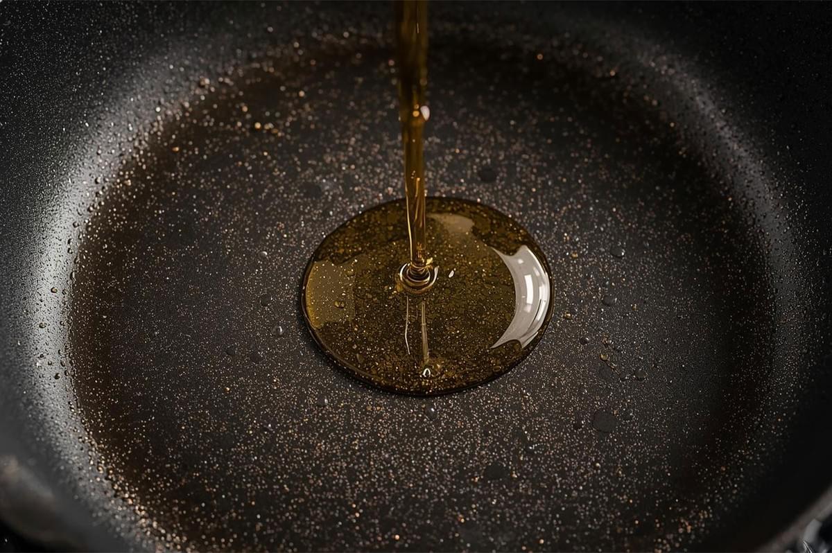 Close-up shot of a steady stream of cooking oil (such as vegetable or grapeseed oil) being poured into the center of a black frying pan for seasoning or cooking.