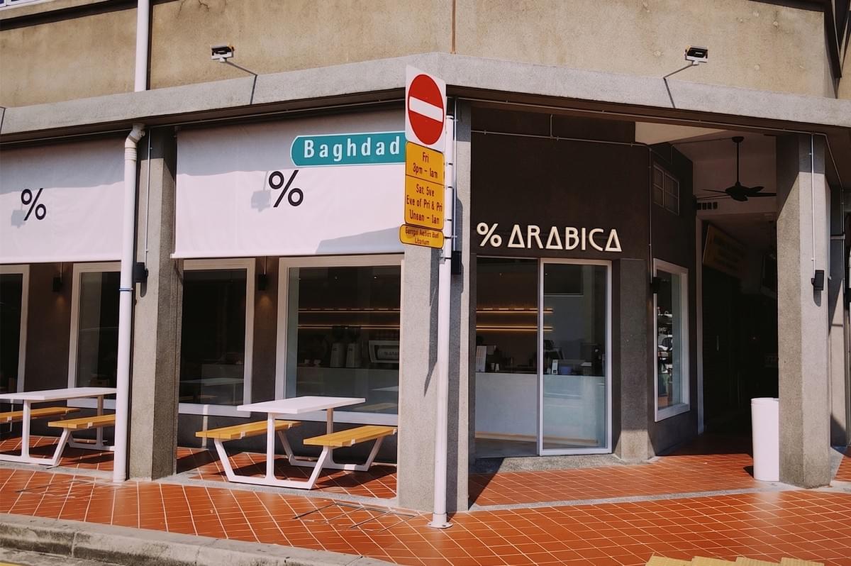 Modern % Arabica coffee shop exterior with outdoor seating and a street sign in a city.