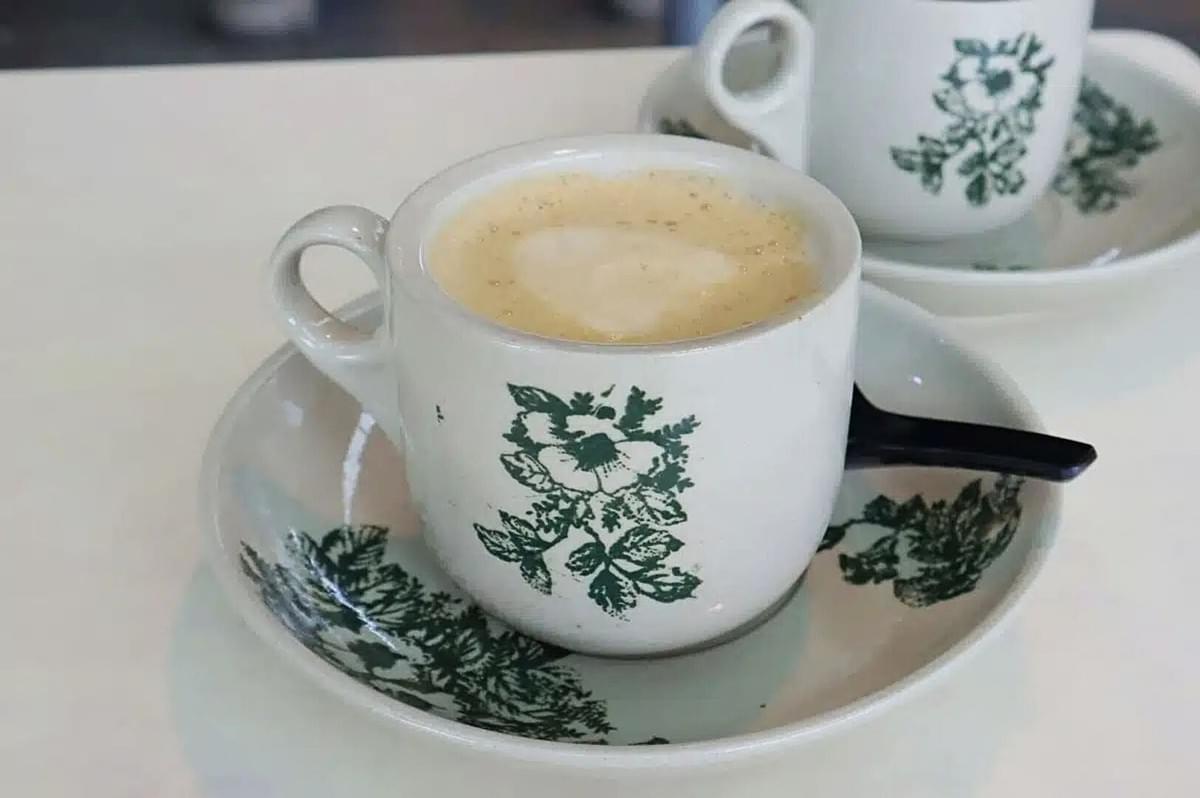 A frothy coffee served in a white cup with green floral designs, placed on a matching saucer with a small black spoon. The tone is cozy and inviting.