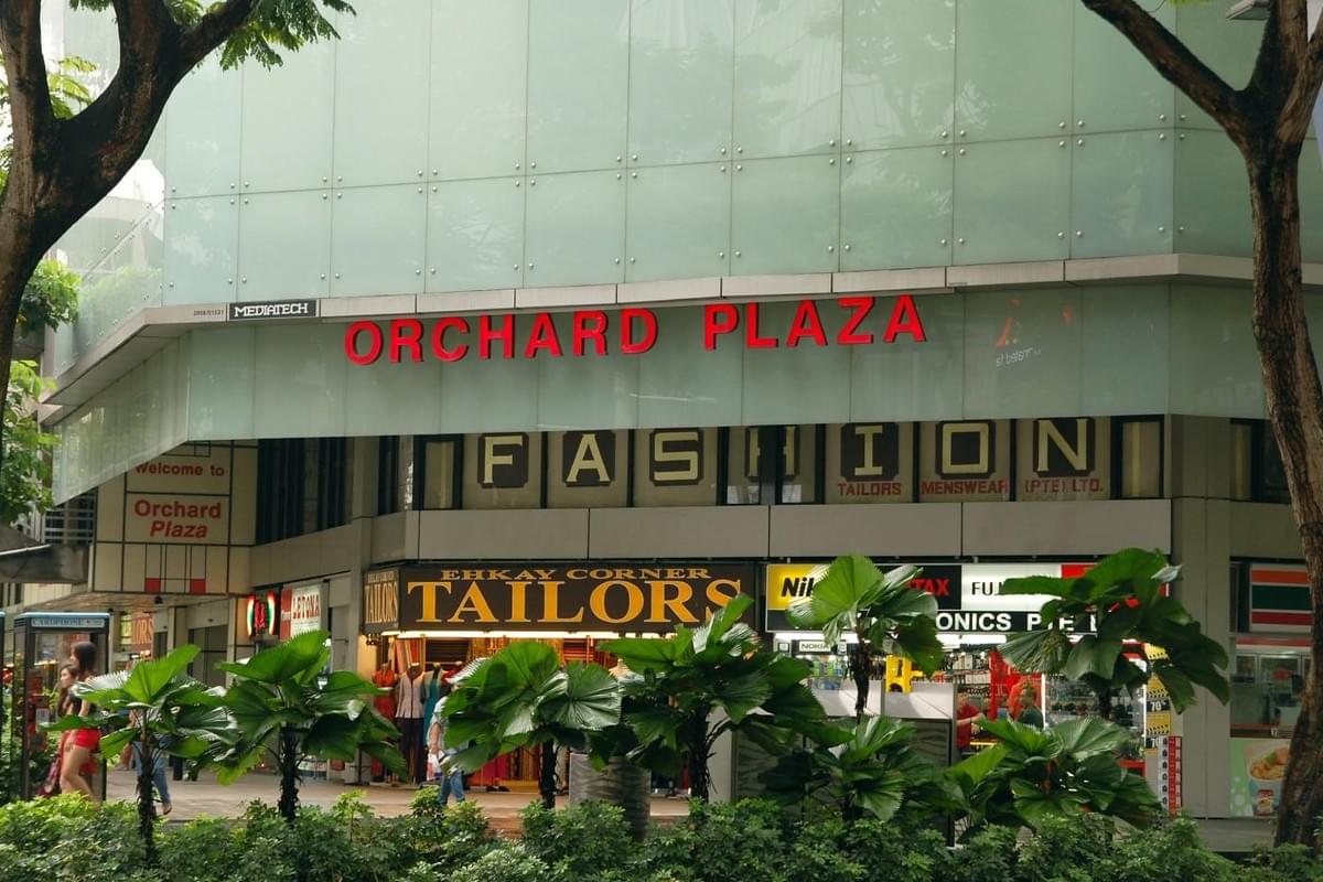 This image captures the exterior of Orchard Plaza, featuring prominent red signage above a storefront for Ehkay Corner Tailors. Lush tropical greenery and mature trees frame the entrance, where people are seen walking past the various shop windows.