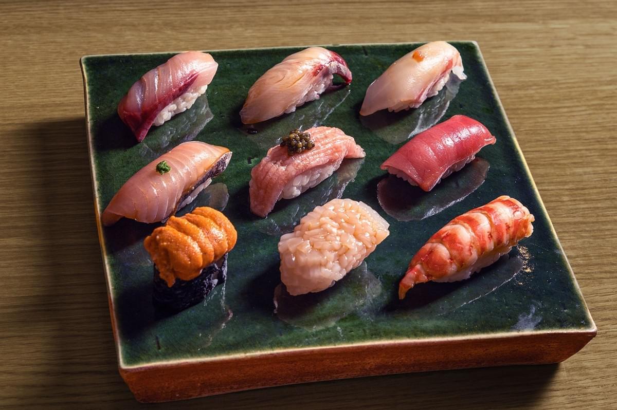 A square green ceramic plate featuring nine pieces of vibrant sushi, including tuna, shrimp, and uni, each artfully arranged in three rows.