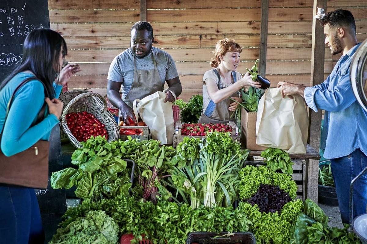 Farmers market stall with people selecting and bagging fresh leafy greens, tomatoes, and other vegetables.