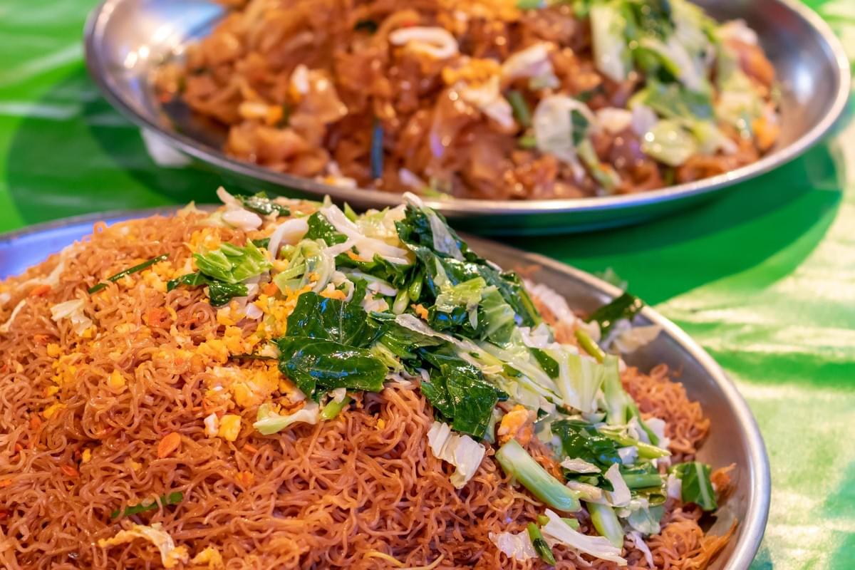 Two large metal platters are piled high with mounds of stir-fried noodles, set against a vibrant green table covering. The dish in the foreground highlights thin, brown vermicelli noodles garnished generously with chopped green vegetables and scattered bits of scrambled egg.