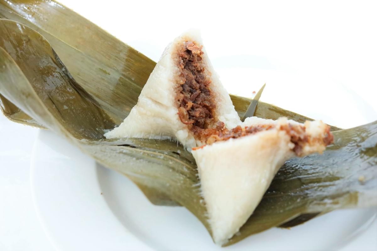 A sticky rice dumpling, known as zongzi, is sliced open to reveal a savory dark meat filling inside. It rests atop a green bamboo leaf on a simple white plate.