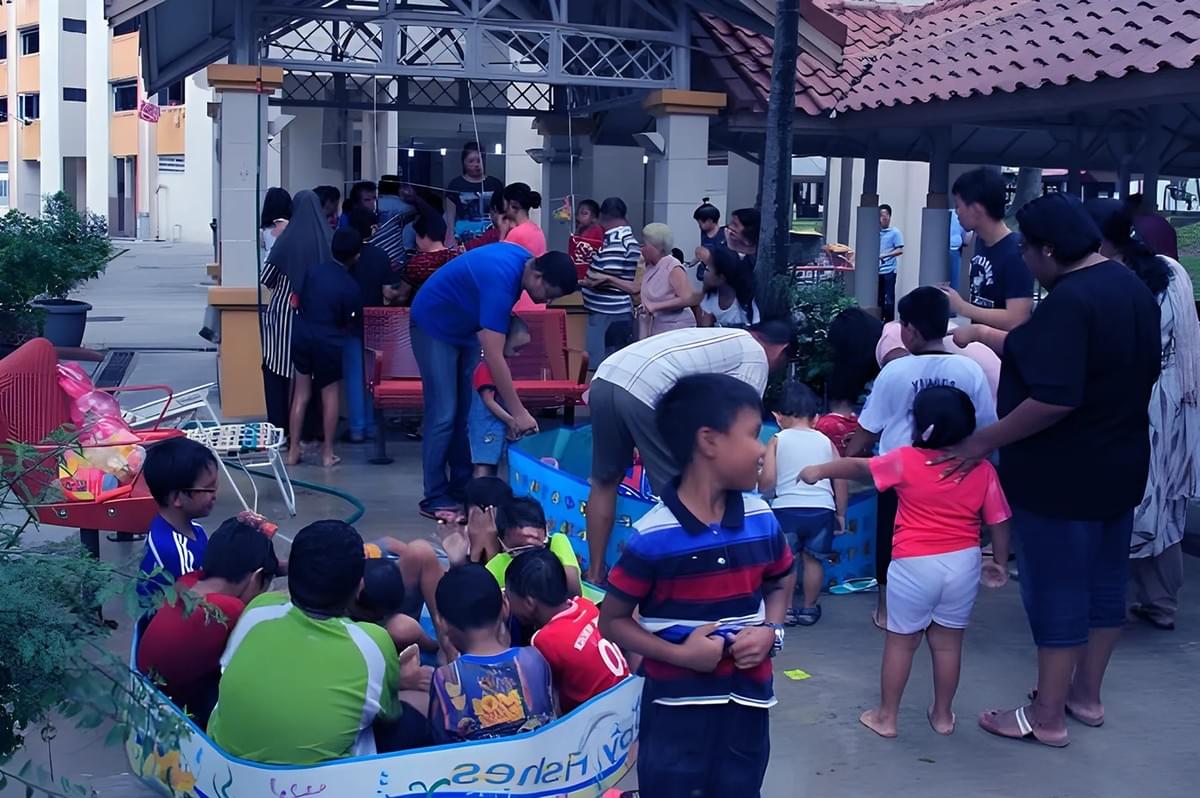 A group of children play in and around a small inflatable pool at a community event. Adults nearby are socializing under a pavilion with a relaxed atmosphere.