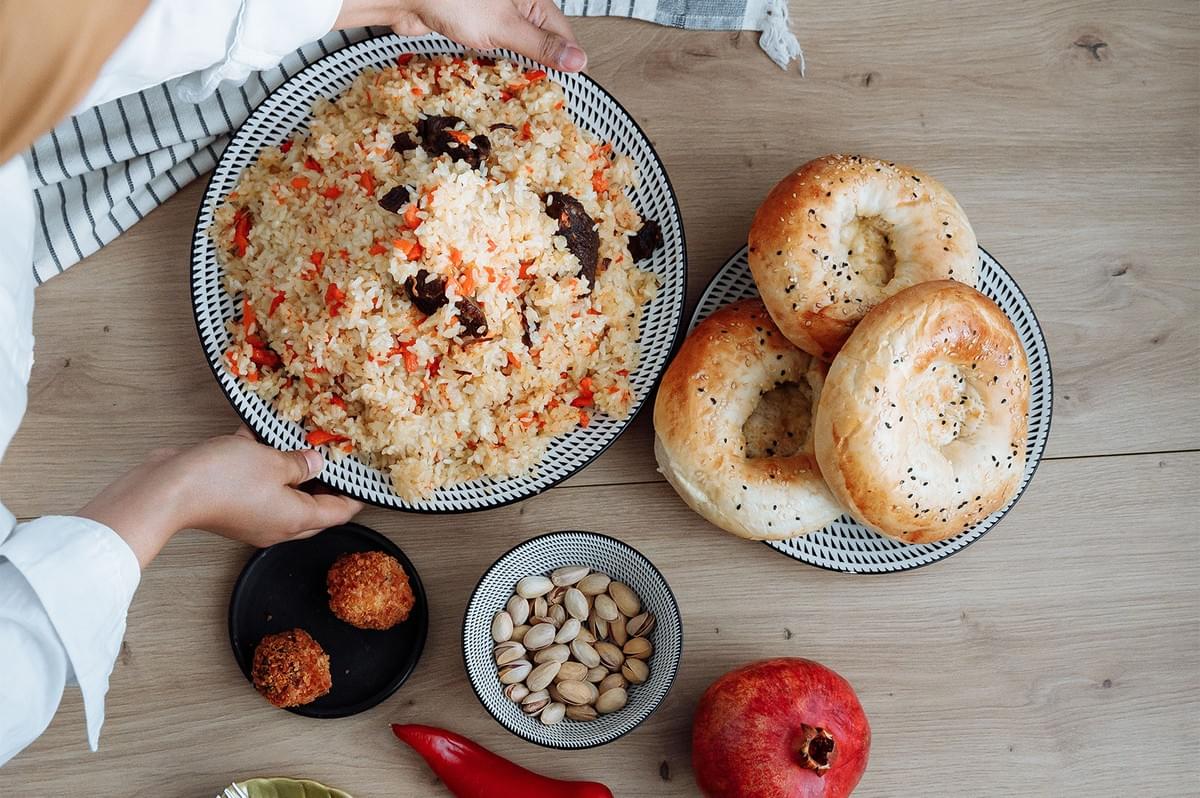 Hands serving halal food spread with rice dish, bread, and condiments on a wooden table.