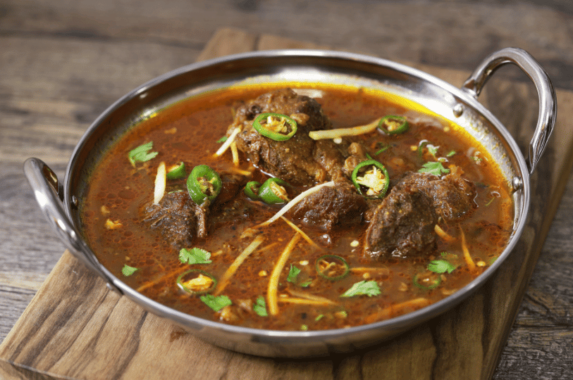 A stainless steel serving dish filled with hearty Pakistani Nihari, featuring deep red spicy gravy garnished with green chilies and ginger.