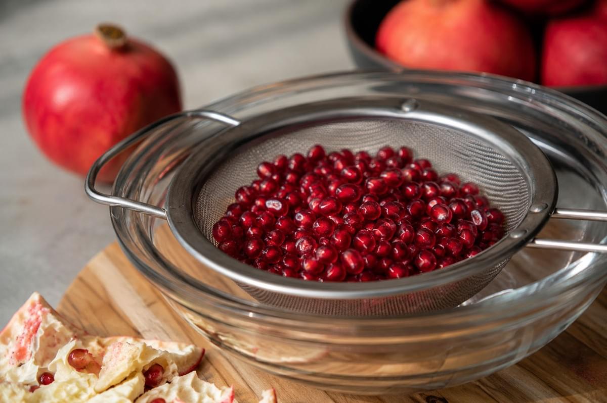 Fresh pomegranate seeds collected in a metal mesh sieve over a glass bowl, with fruit skins and whole pomegranates nearby