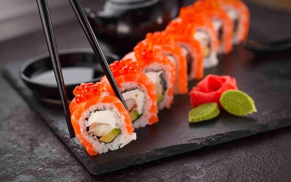 A line of salmon sushi rolls topped with orange fish roe is presented on a dark slate platter alongside wasabi and pickled ginger. Black chopsticks lift a single piece, highlighting its filling of cream cheese, avocado, and fish.